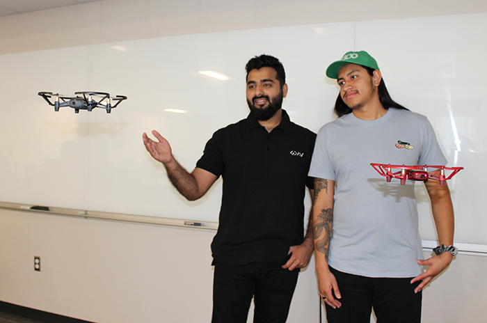 Two computer science students stand next to two hovering drones in a classroom.