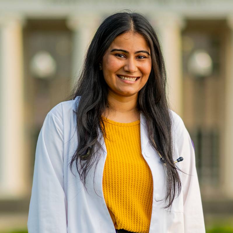 A nursing student with long hair wearing a gold shirt under her white doctor's coat smiles at the camera.